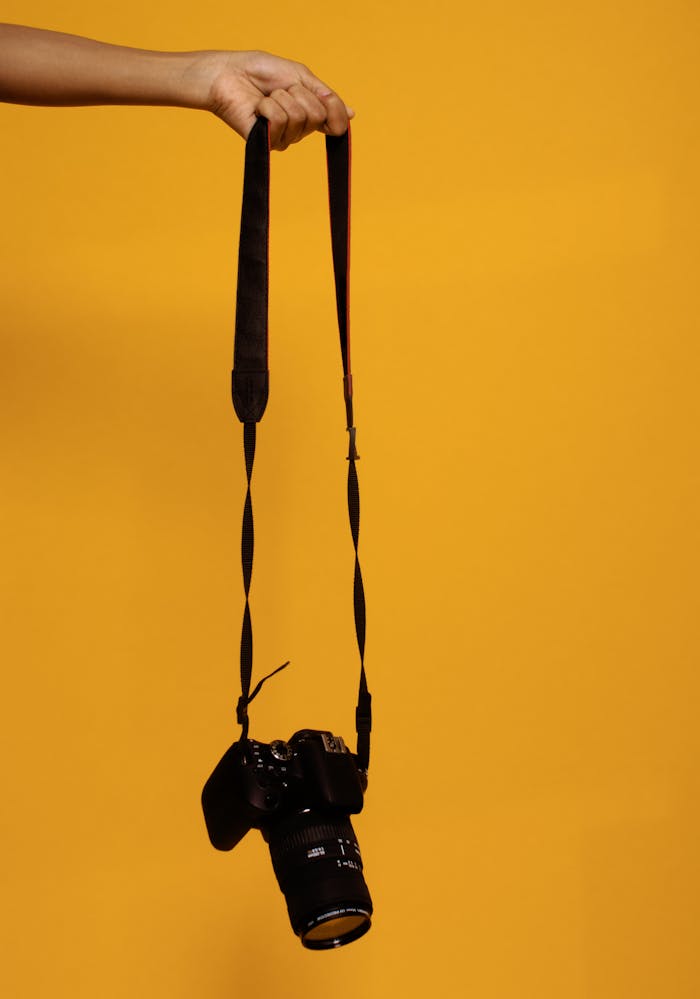 services-04 A DSLR camera suspended by hand against a vibrant orange backdrop, showcasing photography technology.